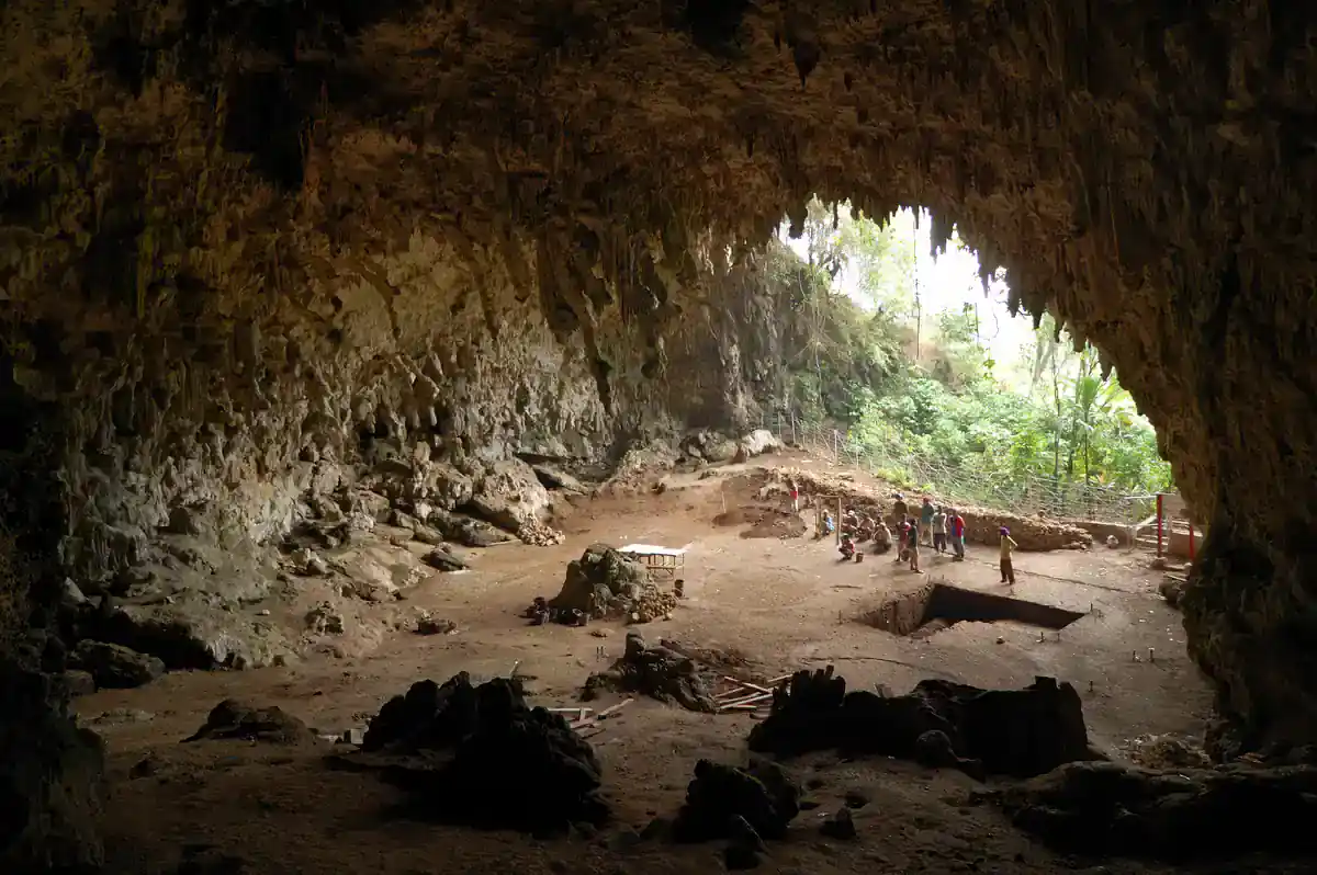 Wide view inside a large limestone cave with an excavation trench on the floor, people working near the bright entrance and forest outside
