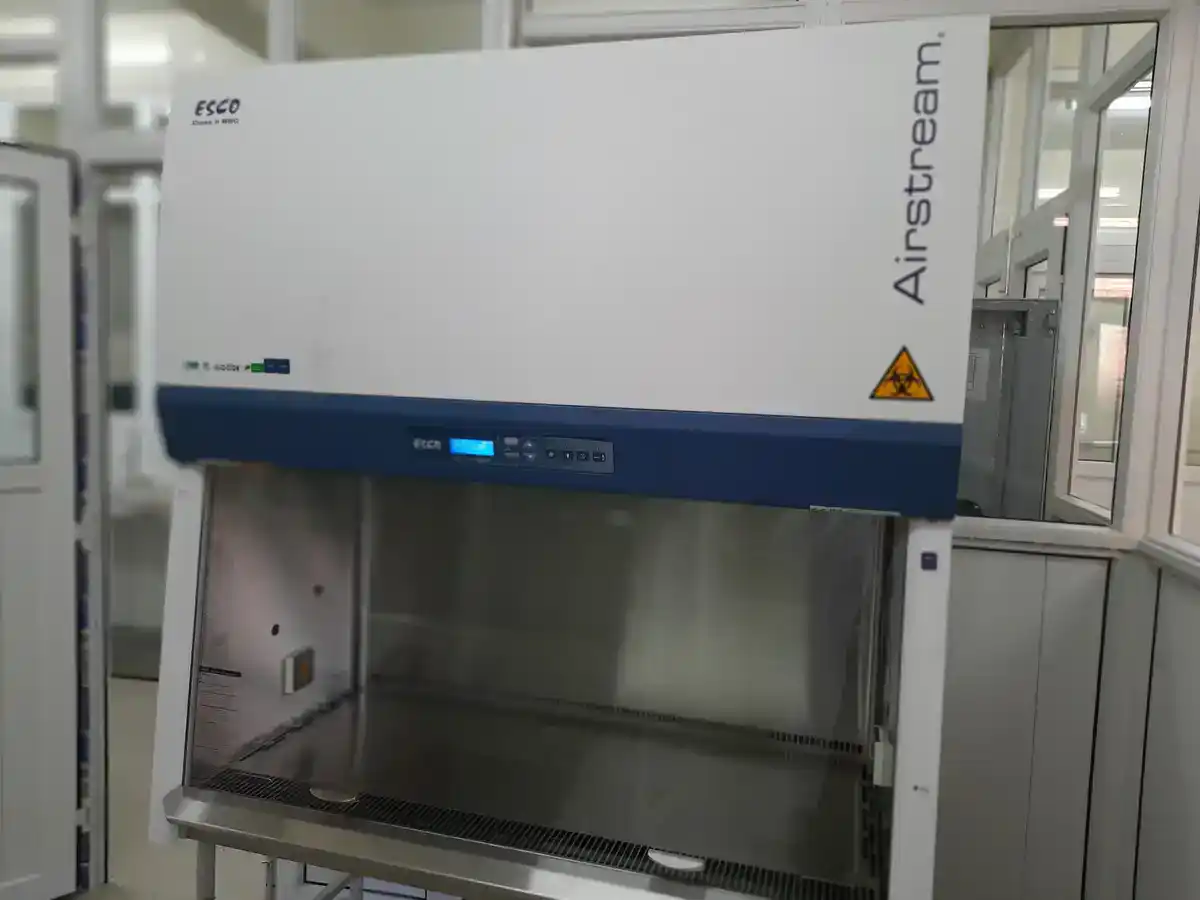 Class II biosafety cabinet in a laboratory, sash raised and work surface empty, with a biohazard symbol on the front panel and a digital control display along the front edge.